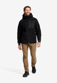 Svart fleecejacka med en zippered bröstficka och strukturerad tyg, kombinerad med tan-byxor och svarta skor, visad i en stående pose.