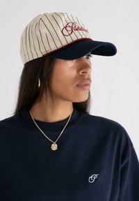 Baseballcap med kremfarget pinstripe-stoff, rød brodert tekst, og en marineblå skygge. Brukt med en marineblå t-skjorte og gullkjede.