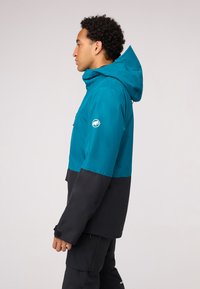 Türaun- und schwarze Jacke mit Kapuze, ausgestattet mit einer Fronttasche, strukturiertem Gewebe und verstellbaren Bündchen. Logo auf dem linken Ärmel sichtbar.