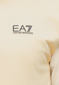 Béžová mikina s rebrovanou textúrou a logom „EA7“ a „EMPORIO ARMANI“ v tmavých farbách na prednej strane.