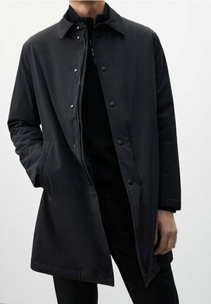 Mężczyzna ubrany w czarny płaszcz o długości do połowy uda z zapięciem na guziki i zamek, stojący z jedną ręką w kieszeni płaszcza na białym tle.