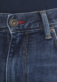 Primer plano de la cintura de unos pantalones de mezclilla azul con botón de Tommy Hilfiger, detalle de costura roja y trabilla para cinturón sobre un fondo de tela oscura.