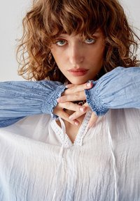 Blusa bianca leggera con una finitura testurizzata e scollatura a V, completata da maniche a sbuffo blu con polsini arricciati.