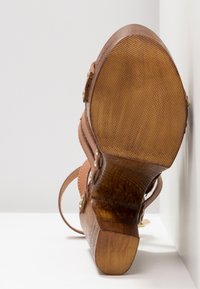 Sandale compensée en cuir marron avec semelle texturée et rivets dorés, présentée semelle vers le haut contre un fond et un mur blancs.