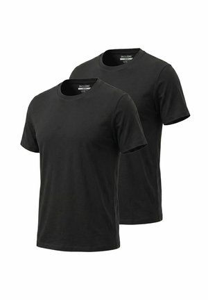 Junger Mann mit lockigem Haar, trägt schwarzes T-Shirt, schwarze Hose, weiße Turnschuhe, steht mit verschränkten Armen und lächelt, Tätowierungen an den Armen sichtbar.