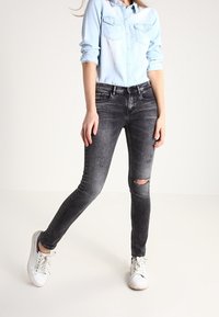 Mörkgrå tajta jeans med ett rivet knä, ihop med en ljusblå skjorta med knappar och vita sneakers.