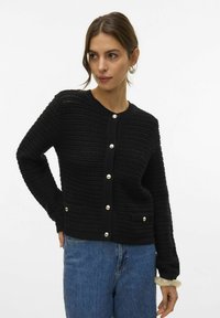 Donna che indossa un cardigan nero a bottone con texture, bottoni argentati e jeans blu, in posa con una mano sui fianchi.