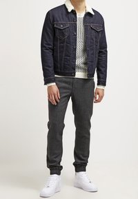 Veste en denim foncé avec un col en polaire blanche, pull blanc à motif, pantalon de jogging gris, et baskets blanches.