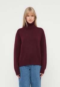 Pulover cu guler înalt burgund, confecționat dintr-un material moale și texturat. Design lejer, cu manșete și tiv ribbate, asortat cu blugi largi de culoare albastră.