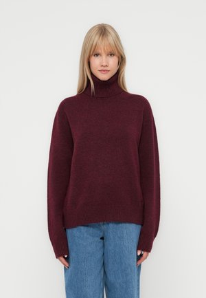 Burgundy turtleneck sweater in soft, textured fabric. Loose-fitting design with ribbed cuffs and hem, paired with blue wide-leg jeans.