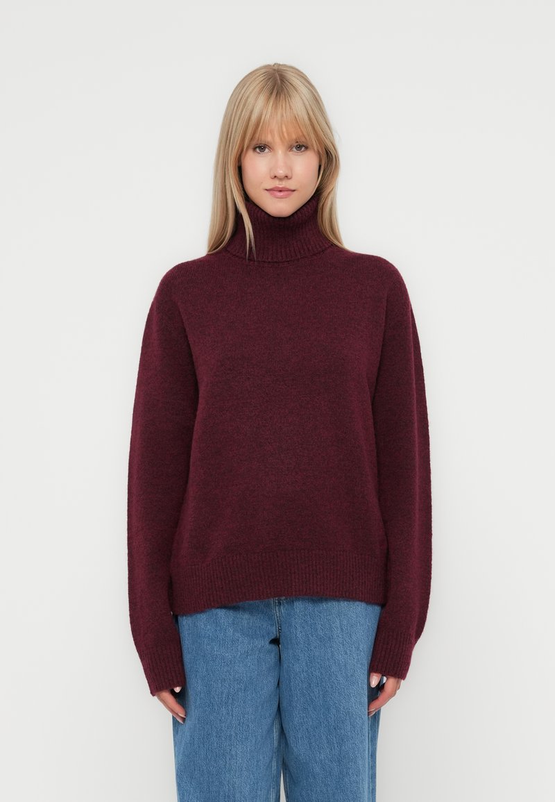 Pulover cu guler înalt burgund, confecționat dintr-un material moale și texturat. Design lejer, cu manșete și tiv ribbate, asortat cu blugi largi de culoare albastră.