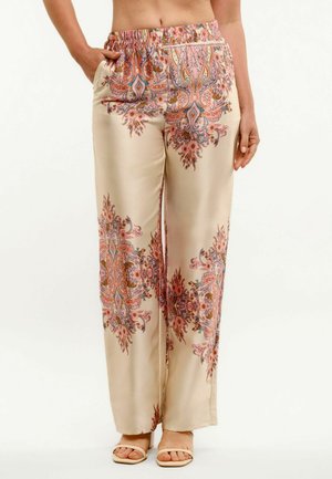 Bež široke hlače s ružičastim i plavim cvjetnim paisley uzorcima, elastičnim strukom, lijeva ruka modela u džepu, uparen s bež sandalama otvorenih prstiju.