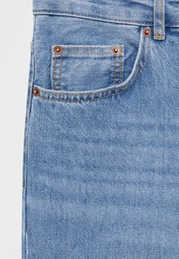 Jean en denim bleu clair en gros plan montrant la poche avant, petite poche à monnaie avec rivets en cuivre et passants de ceinture avec surpiqûres orange.