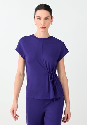 Femme aux cheveux foncés attachés porte un haut violet à manches courtes avec un détail de nœud sur le côté et un pantalon assorti sur un fond clair uni.