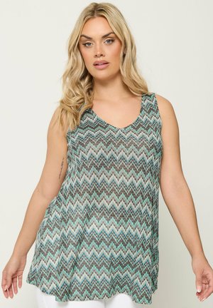Blond vrouw die een mouwloos V-hals top met blauw, zwart en wit zigzagpatroon draagt, staand tegen een effen lichte achtergrond.