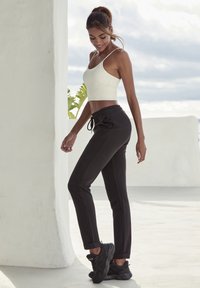 Schwarze Jogginghose mit Kordelzugbund, schmal zulaufenden Beinen und Seitennähten; kombiniert mit einem weißen, taillierten Croptop und schwarzen Sportschuhen.