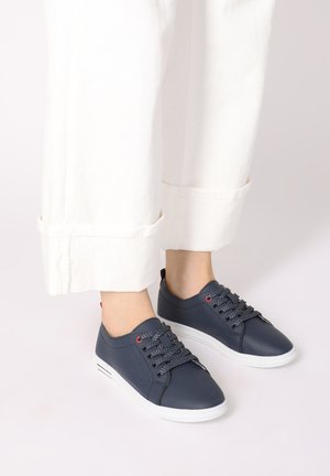 Nachtblaue Leder-Sneaker mit grauen Schnürsenkeln, weißer Sohle und roten Ösenakzenten; kombiniert mit weißen, hochgekrempelten Hosen.