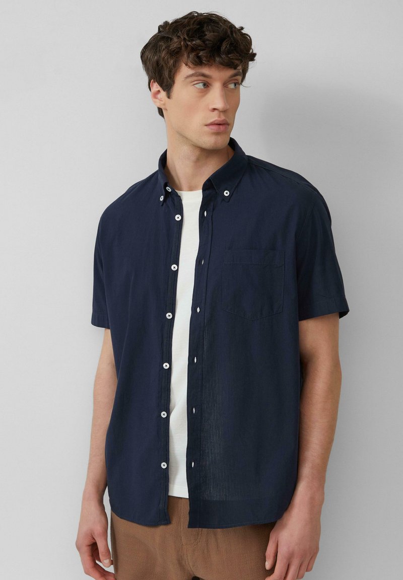 Camisa de botones de manga corta en color marino, hecha de un material ligero, con un bolsillo en el pecho y botones blancos, llevada sobre una camiseta blanca.