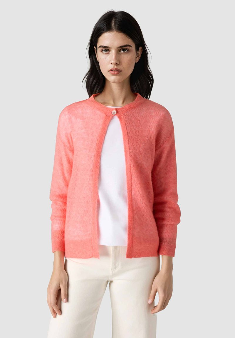 Frau mit dunklen Haaren, die eine korallenrosa flauschige Strickjacke trägt, die oben über einem weißen Hemd und cremefarbenen Hosen zugeknöpft ist, nach vorne blickend.