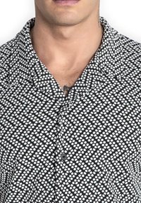 Camisa negra de manga corta con un patrón geométrico de puntos blancos, cierre de botones y cuello clásico. Tejido ligero con textura suave.