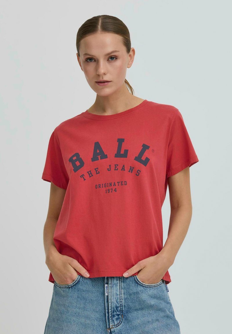 Femme portant un T-shirt rouge avec le texte "BALL THE JEANS ORIGINATED 1974", associé à un jean en denim bleu clair, les mains dans les poches, sur un fond neutre.