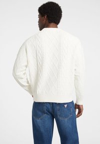 Pullover bianco chunky a maglia con pattern a diamante testurizzati, polsini e orlo a coste. Indossato con jeans in denim blu, visto da dietro.