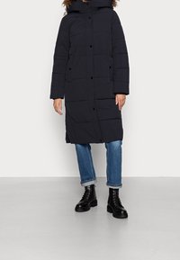 Abrigo acolchado azul marino con capucha, con secciones acolchadas, botones de presión y mangas largas, combinado con vaqueros azules y botas negras con cordones.