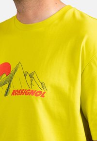 Žluté tričko s krátkým rukávem vyrobené z bavlny, zdobené grafikou hor a nápisem "ROSSIGNOL" tučným červeným písmem.