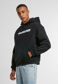 Zwarte hoodie van zacht materiaal, met een voorkantzak en grafische tekst. Gecombineerd met lichtblauwe spijkerbroek.
