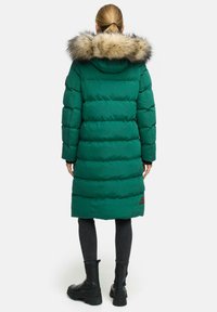 Zeleni napihnjen plašč s kapuco oblečeno z krznom, ki ima vodoravne šive, dolge rokave in srednje dolžino. Črne podrobnosti na robu.