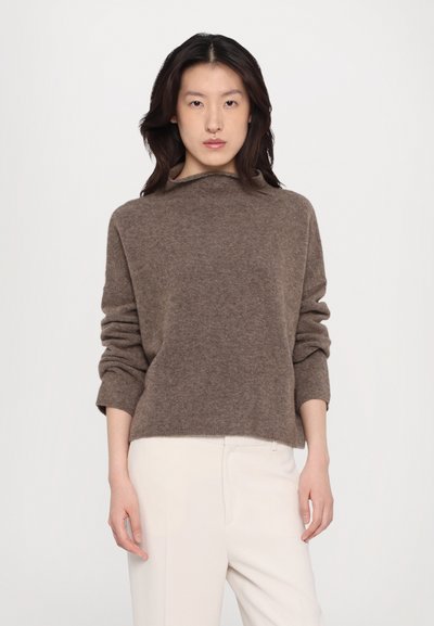 Brun, myk strikkegenser med funnel-neck, løs passform og rullede ermer, sammen med lyse bukser. Enkel og minimal design.