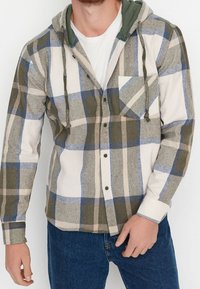 Camicia a scacchi in flanella con cappuccio verde, caratterizzata dai colori grigio, blu e crema. Include una tasca sul petto e chiusure a bottone. Tessuto morbido.