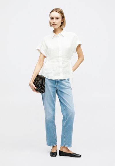 Femme portant un chemisier blanc à manches courtes boutonné, un jean bleu clair, des ballerines noires, tenant un sac à main noir avec des détails en chaîne.