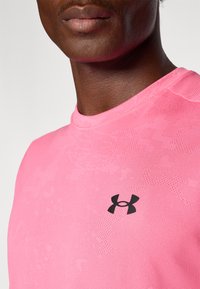 Rozā sporta krekls ar teksturētu rakstu un pieguļošu dizainu, uz krūtīm redzams melns Under Armour logo. Īsas piedurknes, apaļa kakla izgriezums.