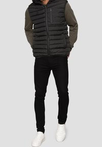 Veste noire matelassée avec capuche, présentant des coutures horizontales et une poche poitrine. Portée sur une chemise à manches longues vert foncé et un pantalon noir avec des baskets.