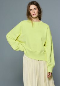 Neonsko rumeno širok pulover z rebrastimi manšetami in robom, kombiniran z lahkotno kremno krilo, postavljen na sivi podlagi.