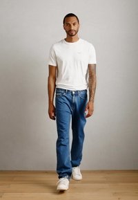 Mann som går innendørs iført en hvit t-skjorte, blå jeans og hvite joggesko, med synlig tatovering på høyre arm mot en enkel grå vegg.