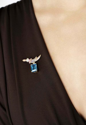 Gold-tone brooch with a central blue rectangular gem, accented by purple and pink stones and a pearl, attached to a dark fabric surface.