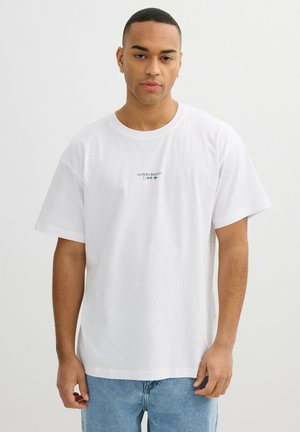 Joven con el pelo corto que lleva una camiseta blanca con texto y símbolos pequeños, combinada con unos vaqueros azul claro, de pie frente a un fondo sencillo.