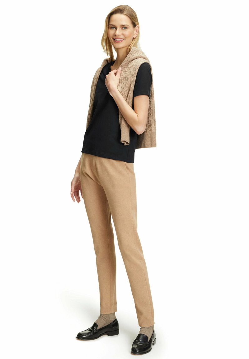 Maglietta nera a maniche corte, gilet beige in maglia drappeggiato sulle spalle, pantaloni beige su misura, calzini grigi e mocassini neri. Design semplice e pulito.