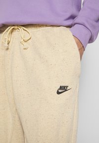 Beiget kirjavat collegehousut, joissa on joustava vyötär�önauha, säädettävä kiristysnauha ja musta Nike-logo vasemmassa taskussa. Yhdistetty violettiin yläosaan.