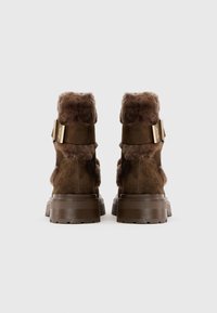 Par de botas de ante marrón con forro de piel gruesa, suelas gruesas y correas con hebilla dorada, mostradas desde atrás sobre un fondo liso.