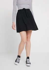 Kvinna som bär en svart kjol med knytbart midjeband, randig långärmad topp och svarta högskor, stående mot en ljus vit bakgrund.