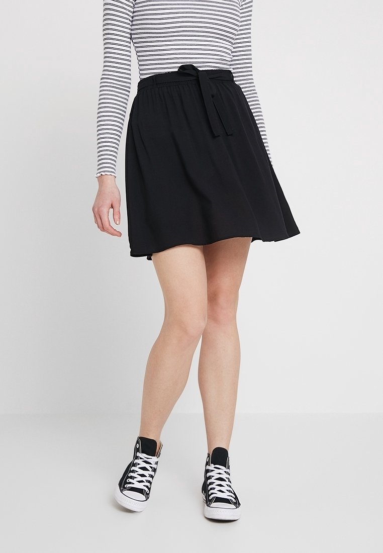 Kvinna som bär en svart kjol med knytbart midjeband, randig långärmad topp och svarta högskor, stående mot en ljus vit bakgrund.