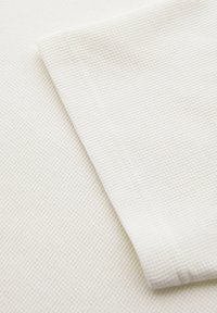 Dva překrývající se kusy bílé látky s texturou vafle, s viditelným šitím na okraji horního kusu.