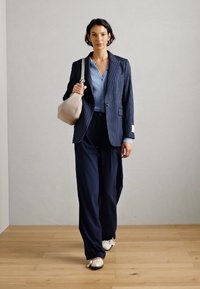 Marineblau gestreifter Blazer, hellblaues Hemd, weite marineblaue Hose, weiße Sneaker und eine beigen Umhängetasche vor einem neutralen Hintergrund.