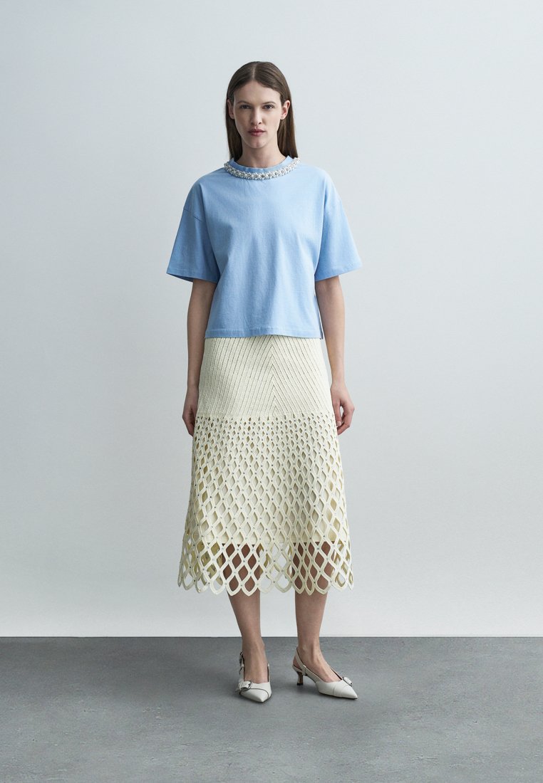 Femme portant un haut à manches courtes bleu clair avec un col orné et une jupe midi couleur crème à motif ajouré, debout sur un sol gris.