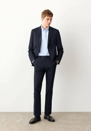 Mornarsko plavo odijelo s uskim sakoom i krojenim hlačama, u kombinaciji sa svijetloplavom košuljom i crnim mokasinama, na jednobojnoj pozadini.