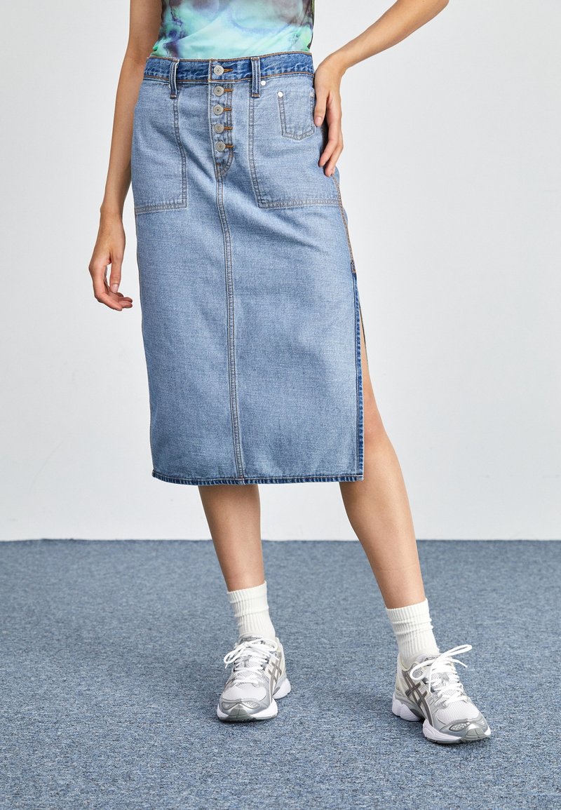 Jupe en denim bleu clair avec une coupe droite, fermeture à boutons sur le devant et fente latérale. Portée avec des chaussettes blanches et des chaussures de sport grises.