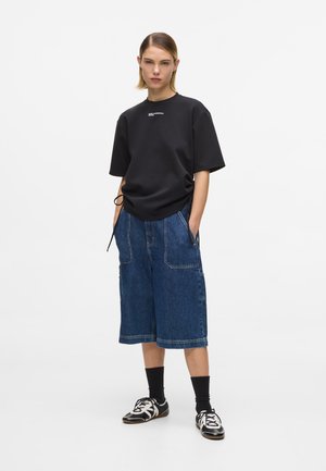 Tânăr în picioare cu mâinile în buzunare, purtând un tricou negru supradimensionat, pantaloni scurți de denim largi până la genunchi, șosete negre și adidași negri cu alb.